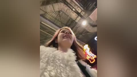 Media: Video of a young woman with light skin and long, light brown hair, wearing a white fluffy jacket, smiling, standing in an industrial, dimly lit room with exposed metal beams and colorful neon lights in the background.