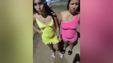 Media: Video of two young women, one in a yellow dress and the other in a pink one-piece, standing closely together on a dimly lit street.