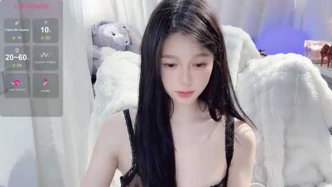 Media: Video of a young Asian woman with long black hair, fair skin, wearing a black lace bra, sitting on a white fur-covered couch in a cozy, soft-focused room.