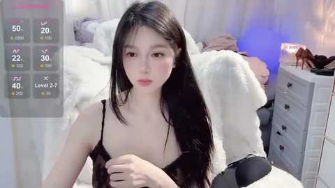 Media: Video of a young Asian woman with long black hair and pale skin, wearing a black spaghetti-strap dress, sitting on a bed in a dimly lit room with a white fur blanket.