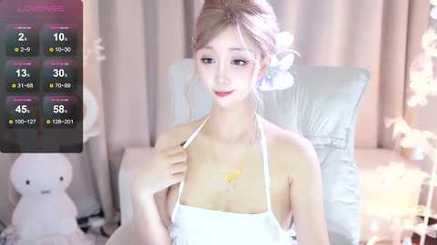 Media: Video of a slender, fair-skinned Asian woman with light brown hair styled in a bun, wearing a white halter top, seated in a cozy, softly lit room with white curtains and plush toys.