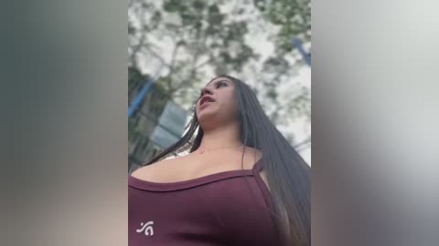 Media: A video of a young woman with long, straight dark hair, wearing a maroon tank top, standing in an urban setting with blurred trees in the background.