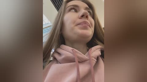 Media: A video of a young Caucasian woman with light brown hair, wearing a pink hoodie, gazing upward with a thoughtful expression. The background includes a partially blurred, beige wall and a blue patterned item.