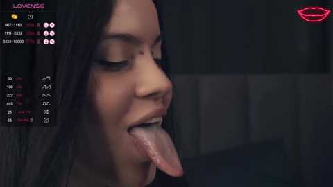 Media: A close-up video of a young woman with dark hair, eyes closed, and long tongue extended. The background is dimly lit, with a smartphone screen displaying \"LOVE ME\" at the top.