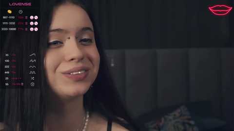 Media: Video of a smiling woman with long black hair, wearing a black top, in a dimly lit room with a bed and a headboard in the background.