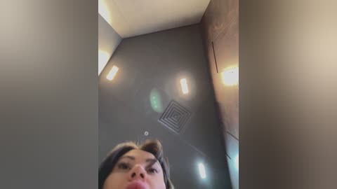 Media: A video of a woman with brown hair, captured from a low angle, standing in a dimly lit, narrow hallway with beige walls and a rectangular ceiling light.