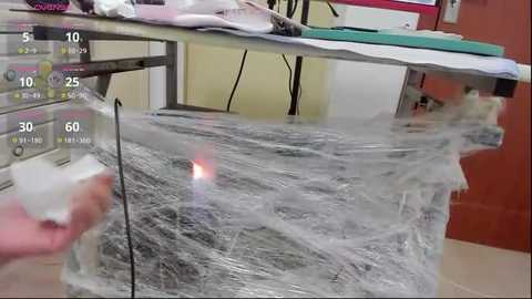 Media: Video of a medical procedure in a sterile room, showing a person's hand applying a white plastic wrap to a patient's chest. Medical equipment and a monitor displaying vital signs are in the background.