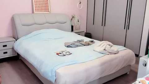 Media: Video of a modern, minimalist bedroom with a light gray upholstered bed, light blue bedspread, and cream throw blanket. Pastel pink walls, white nightstands, and built-in wardrobe with vertical handles.