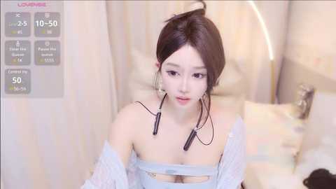 Media: Video of a young Asian woman with fair skin, dark hair, and light makeup, wearing an off-shoulder white lace dress, sitting on a bed with a furry blanket, in a soft-lit, pastel-colored room.