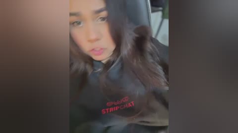 Media: Video of a young woman with long dark hair, wearing a black \"Stripper\" hoodie, sitting in a car, with a blurred background.