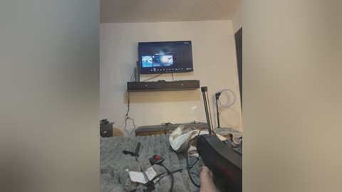 Media: Video of a cluttered, dimly-lit bedroom with a TV mounted on a wall, a bed with grey blankets, and a person holding a remote control, surrounded by scattered electronics.