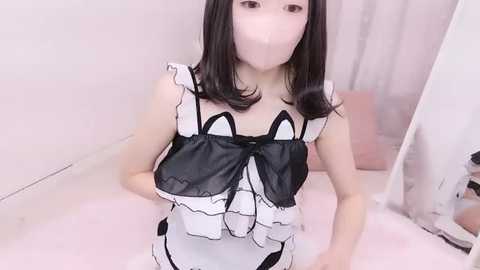 Media: A young East Asian woman with fair skin and shoulder-length black hair, wearing a revealing black and white French maid outfit, standing in a softly lit room with pink decor.