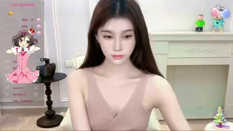 Media: Video of an East Asian woman with long black hair, wearing a pink sleeveless dress, seated indoors. Background features a calendar, dollhouse, and decorative items.