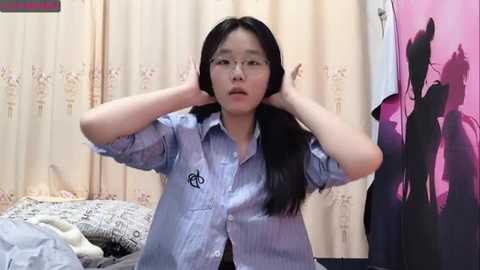 Media: Video of a young Asian woman with long black hair, wearing glasses, adjusting her hair in front of a bed with patterned sheets. Background includes beige curtains and a pink t-shirt hanging on the right.