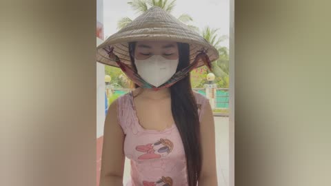 Media: Video of a young woman with medium skin tone and long black hair, wearing a pink tank top with cartoon characters, a traditional conical hat, and a white face mask, standing outdoors in a tropical garden with palm trees.