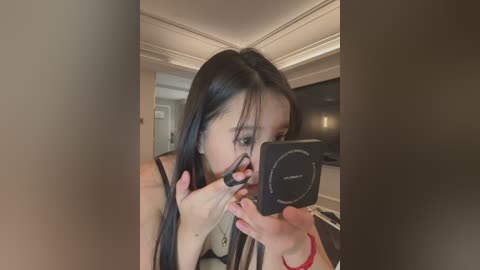 Media: A video of a young East Asian woman with long black hair, wearing a black top, taking a selfie in a modern, dimly lit room with cream-colored walls and recessed lighting.