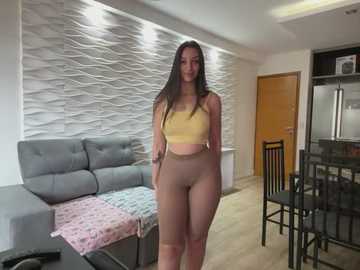 Media: Video of a slender, light-skinned woman with long brown hair, wearing a yellow crop top and beige leggings, standing in a modern living room with a gray couch, wooden door, and wavy white wall art.