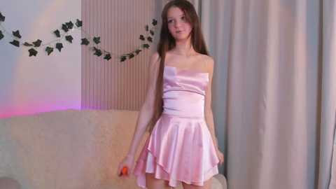 Media: Video of a slender, fair-skinned young woman with long brown hair, wearing a strapless pink satin dress, standing in a softly lit room with floral wallpaper and draped curtains.