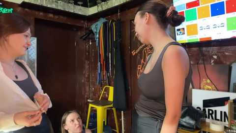 Media: Video of three women in a rustic bar, one in a gray tank top and jeans, another in a black top and white jacket, and a third with glasses, chatting. Background includes a TV, colorful signs, and hanging jackets.