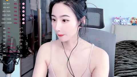 Media: Video of an Asian woman with fair skin, dark hair, and red lipstick, wearing a beige camisole, seated in a room with a grey chair, pink microphone, and plush toys on a shelf in the background.