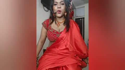 Media: Video of a South Asian woman with medium skin tone, wearing a red saree, adorned with gold jewelry, including a choker, earrings, and bracelets, posing provocatively in a dimly lit room.