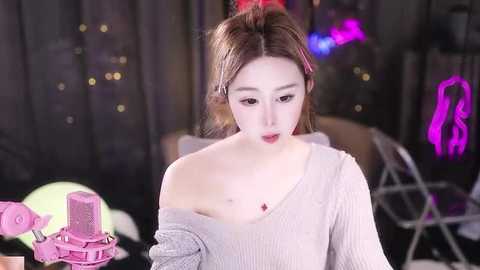 Media: Video of a young Asian woman with fair skin, wearing a light grey off-shoulder sweater, recording herself on a pink microphone stand in a dimly lit, neon-lit room.