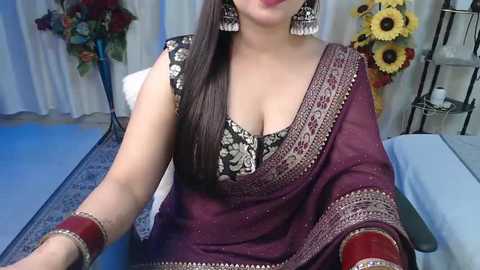 Media: Video of a woman with light skin and long, straight dark hair, wearing a maroon sari with intricate silver embroidery, a black blouse with floral patterns, and large silver earrings. Background includes floral arrangements, sunflowers, and a massage table.