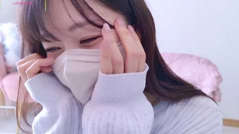 Media: Video of an East Asian woman with straight, shoulder-length brown hair wearing a white surgical mask, looking distressed, with a light pink background.