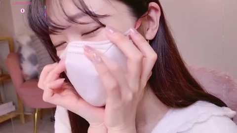 Media: Video of an Asian woman with long black hair, wearing a white face mask, holding it close to her face, sitting on a pink couch in a softly lit room.