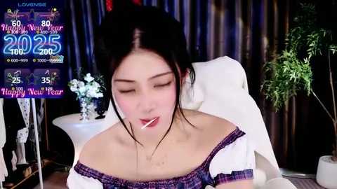 Media: Video of a young Asian woman with fair skin, black hair, and closed eyes, wearing a white off-shoulder top, sitting in a plush chair. Background features festive decorations and a \"2025\" countdown screen.