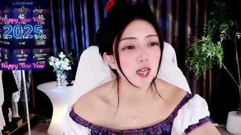 Media: Video of an Asian woman with fair skin, dark hair in a bun, wearing a white off-shoulder dress with purple accents, sitting in front of a TV displaying \"2023\" and \"Happy New Year.\" Background features a potted plant and festive decorations.