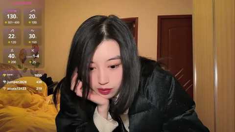 Media: Video of an East Asian woman with shoulder-length black hair, wearing a black jacket and red lipstick, sitting on a bed with yellow bedding. Background shows a beige wall and wooden door.