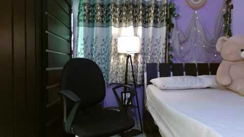 Media: Video of a cozy bedroom with a purple and white color scheme, featuring a bed with plush pillows, a black chair, a lamp, and sheer curtains.