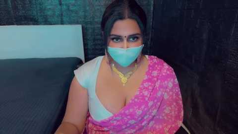 Media: Video of a South Asian woman in a pink saree with white floral patterns, wearing a mask, sitting on a dark bed, surrounded by dark, textured walls.