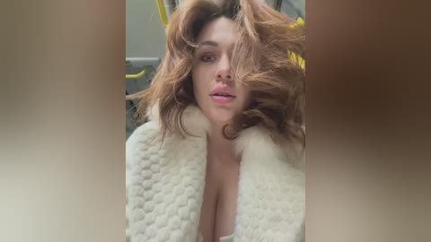 Media: A video of a fair-skinned woman with voluminous, wavy brown hair, wearing a cream-colored, fluffy robe, with a soft, warm indoor setting and a yellow chair in the background.