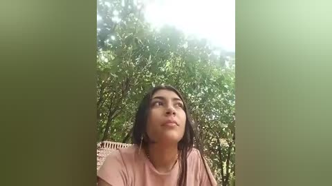 Media: Video of a young woman with medium skin tone, long dark hair, wearing a pink shirt, looking up with a thoughtful expression, surrounded by dense green foliage.