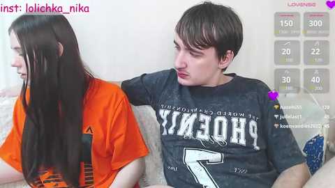 Media: Video of a young woman with long, straight hair in an orange shirt and a young man with short hair in a dark shirt, sitting on a couch, in a dimly lit room.