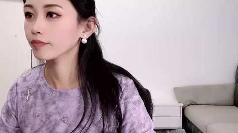 Media: Video of an East Asian woman with long black hair, wearing a lavender patterned blouse, white earrings, and a contemplative expression. Background features a light-colored couch and white wall.