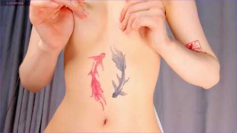 Media: Video of a fair-skinned person with a tattoo of two fish, one red and one gray, on their abdomen. They are topless, with hands raised near their face.