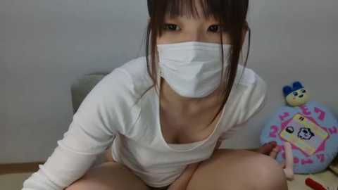 Media: Video of an Asian woman with brown hair, wearing a white mask and long-sleeve shirt, kneeling on a bed with stuffed toys, in a simple room with a plain wall.