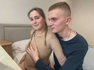 Media: Video of a young couple, a blonde woman with braided hair in a beige top, and a blonde man with a shaved head in a navy shirt, cuddling in a beige bedroom.