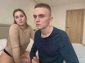 Media: Video of a young couple in a bedroom: a blonde woman in a beige top and black shorts, and a man with short hair in a navy sweater, both looking at the camera.