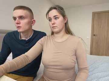 Media: Video of a young, light-skinned couple in casual clothes; man in dark shirt, woman in beige top, seated on a bed in a plain bedroom.