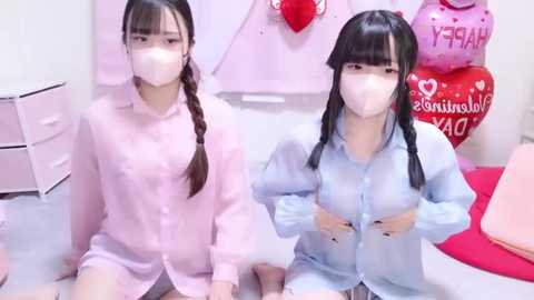 Media: Video of two Asian girls in pastel pink pajamas, wearing white masks, sitting on a carpeted floor in a pink-themed room.