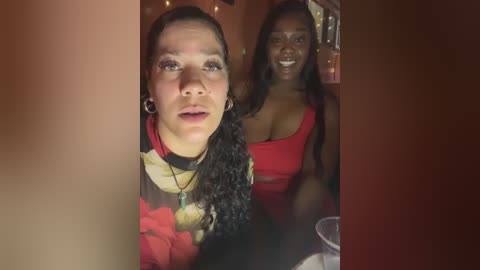 Media: Video of two women with medium skin tone, one with curly hair in red and black floral top, the other with straight hair in red top, smiling, indoors.