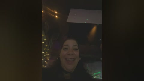 Media: Video of a smiling woman in a dimly lit room, adorned with fairy lights and a projector screen. She has long hair, a black top, and is indoors.