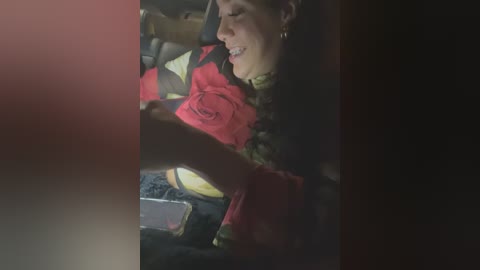 Media: A video captures a smiling woman with short hair, wearing a red and black jacket, holding a red rose. She is seated in a car with dim lighting, her face partially obscured by the frame.