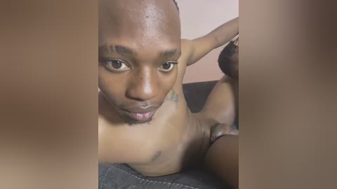 Media: A video of a nude Black man with a shaved head and muscular build, lying on a gray couch, his genitals partially visible. The background is a plain wall.