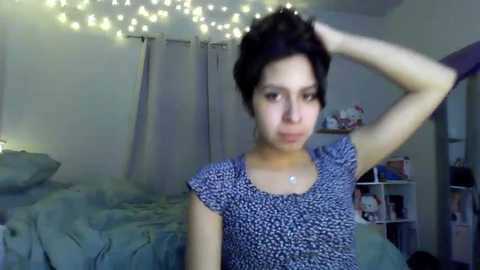 Media: Video of a young woman with short black hair, light skin, and a small build, wearing a blue floral top, standing in a dimly lit bedroom with a bed, white walls, and string lights.