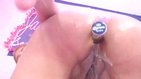 Media: A close-up video of a light-skinned woman's bare buttocks, featuring a gold anal plug branded with the word \"Corona\" and a pink floral arrangement in the background.